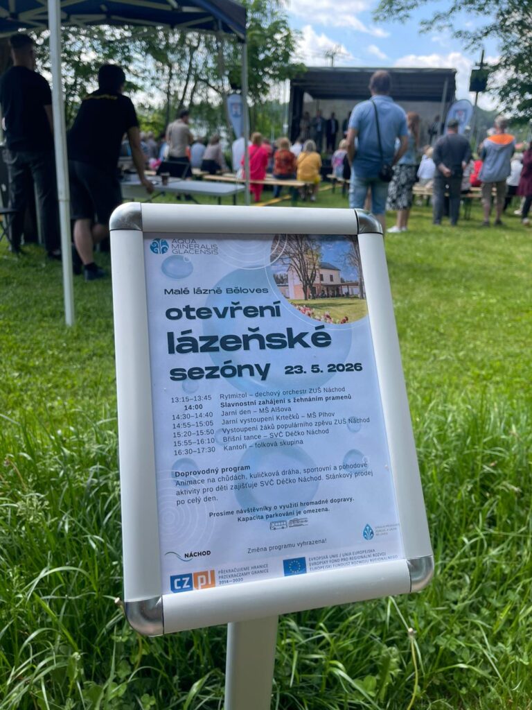 Srebrny potykacz z plakatem programu otwarcia sezonu uzdrowiskowego 2026 na tle zielonej trawy i zgromadzonych ludzi.