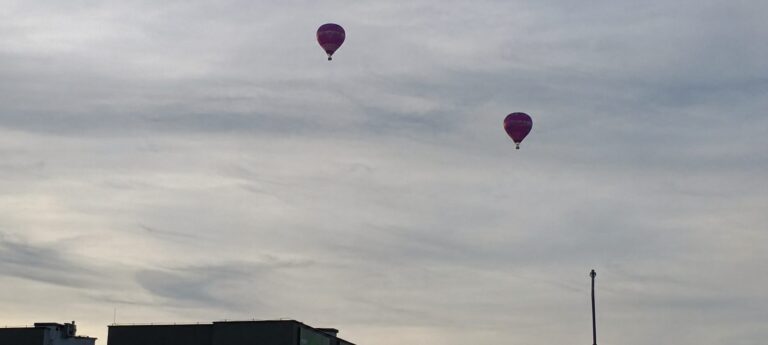 Dwa balony na ogrzane powietrze unoszące się na tle zachmurzonego nieba nad sylwetkami dachów w Kudowie-Zdroju.
