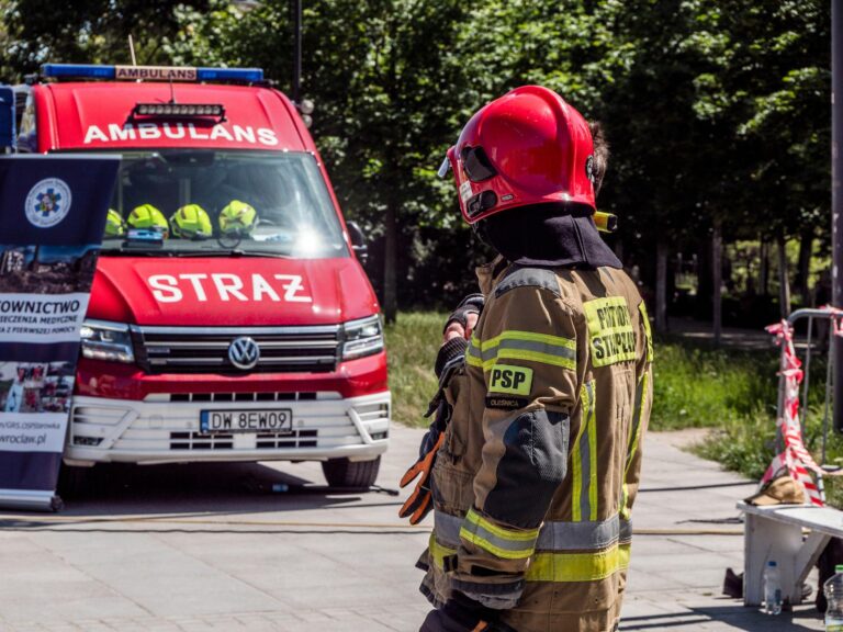 Strażak w pełnym wyposażeniu i czerwonym hełmie podczas akcji ratunkowej, w tle pojazd straży pożarnej i karetka