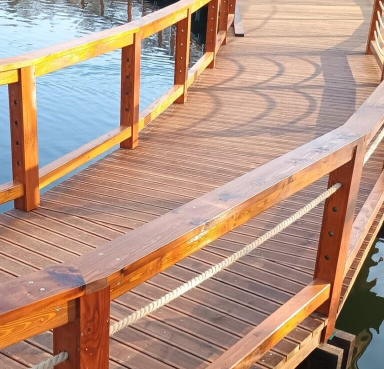Drewniana promenada nad wodą z brązowymi barierkami i linami, rzucająca cienie na deski pomostu.