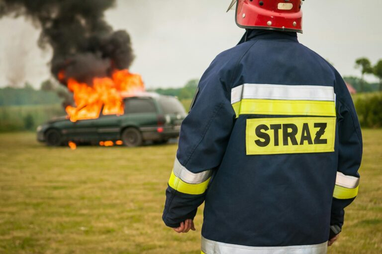 Strażak w granatowym mundurze z napisem STRAŻ i czerwonym kasku stoi tyłem przed płonącym samochodem na polu.