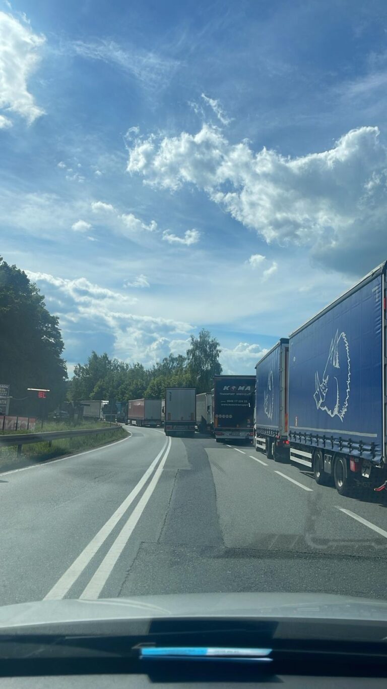 Długi sznur niebieskich i białych ciężarówek stojących w korku na drodze prowadzącej do granicy pod błękitnym niebem.