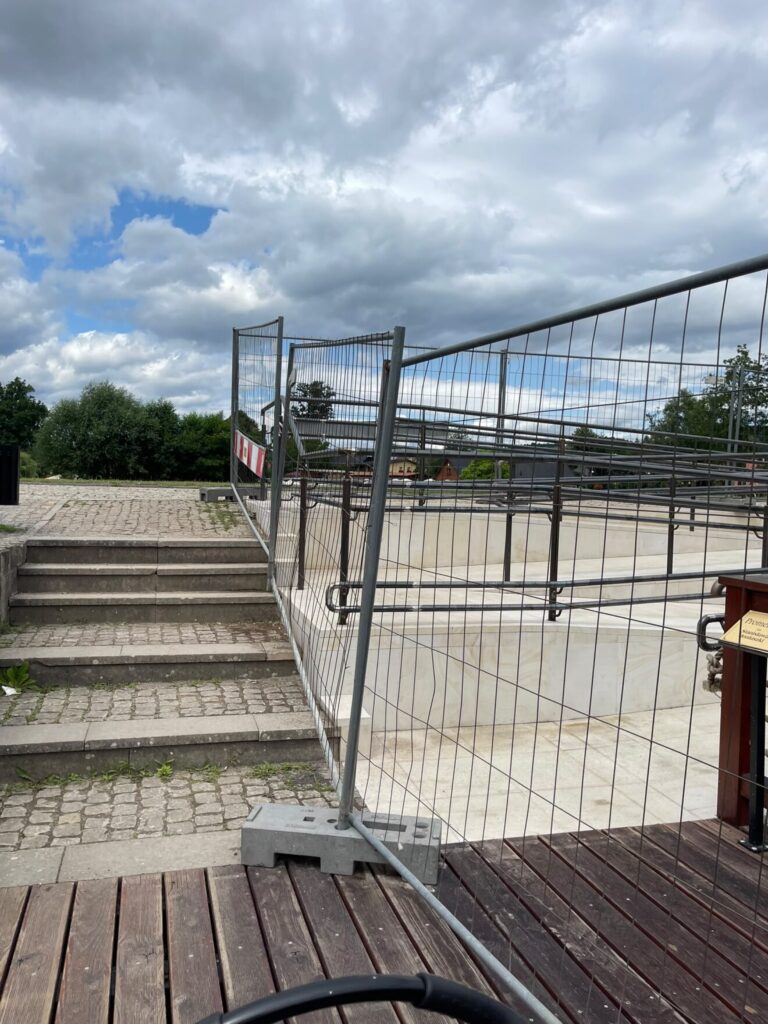 Tymczasowe ogrodzenie budowlane na promenadzie obok kamiennych schodów i jasnej rampy pod zachmurzonym niebem.