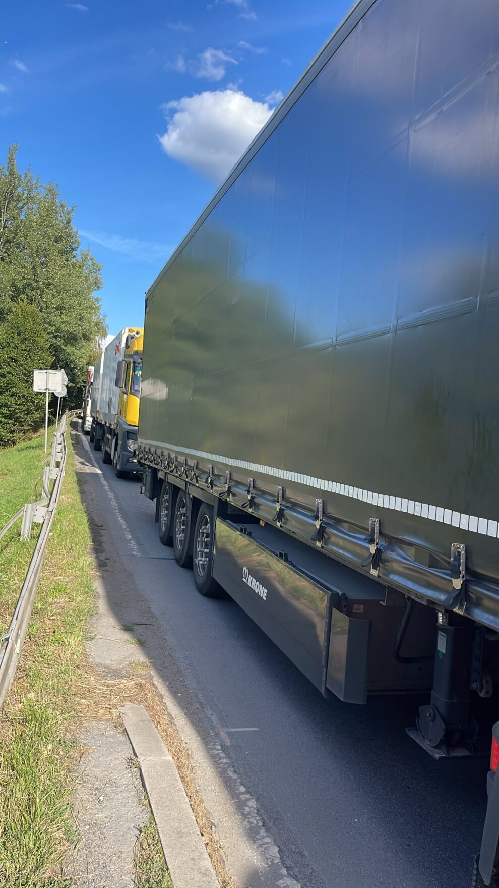Kolejka ciężarówek stojących w korku na wąskiej drodze przy barierach energochłonnych pod błękitnym niebem.