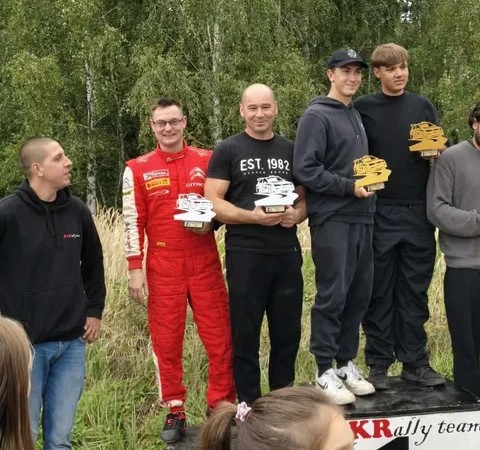 Pięciu mężczyzn na podium, jeden w czerwonym kombinezonie rajdowym, trzymają trofea w kształcie samochodów rajdowych.